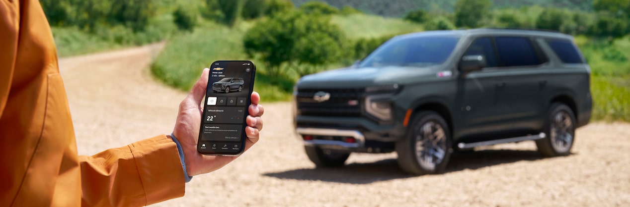Un homme debout dans un paysage montagneux et robuste peut tenir un téléphone intelligent noir affichant l’application mobile Chevrolet avec des commandes à distance du véhicule, avec un Tahoe 2026 bleu visible devant lui.