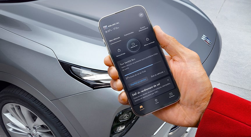 La main d'une personne tient un smartphone noir affichant l'application mobile myGMC montrant les fonctions de démarrage à distance et de climatisation du véhicule sur un fond flou avec des structures rouges et blanches.