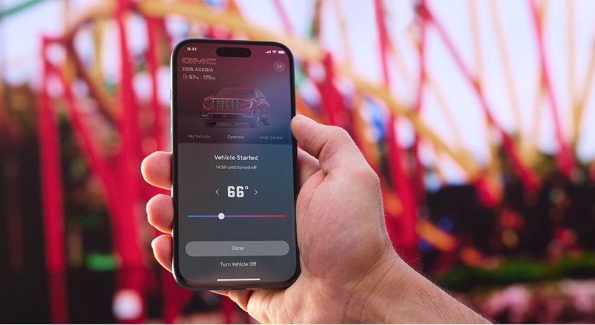A person's hand holds a black smartphone displaying the myGMC mobile app showing the vehicle's remote start and climate control features against a blurred background with red and white structures.