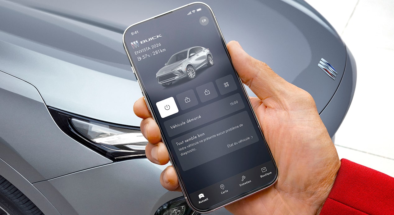 Une main tient un téléphone intelligent affichant l’application mobile Buick avec des commandes à distance du véhicule avec un Envista 2026 gris en arrière-plan d’un environnement extérieur.