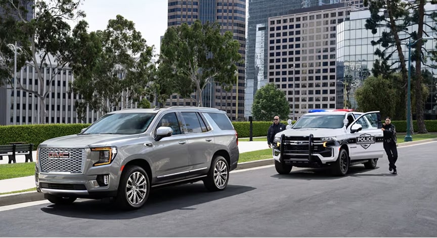 A silver GMC SUV drives on a city street while police officers attend to a scene with a police car in the background.