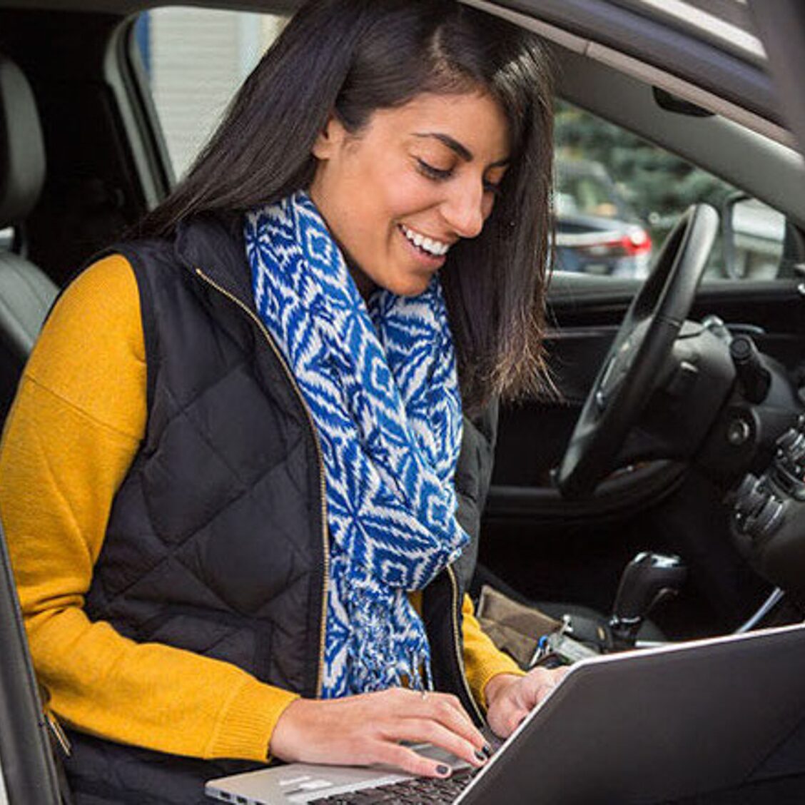 A smiling woman using a laptop to talk to an OnStar advisor while inside her vehicle.