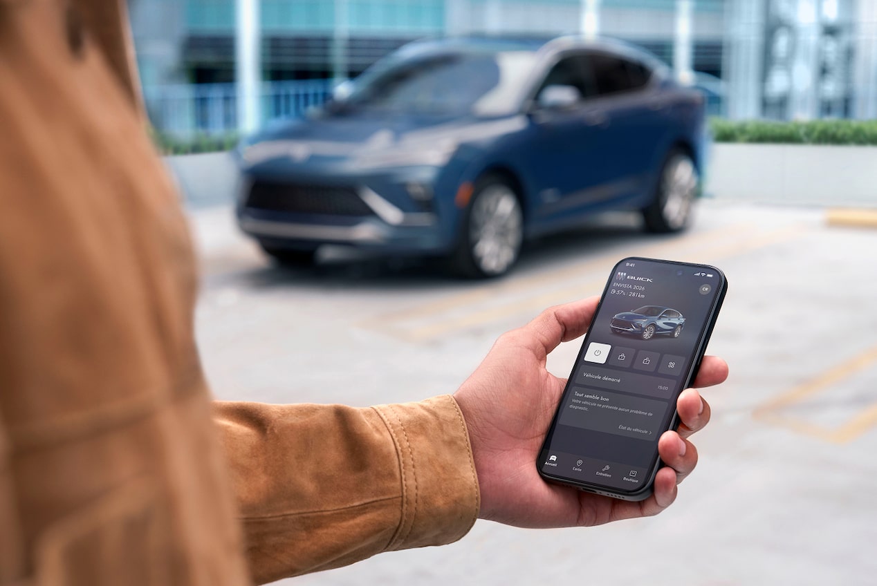 Une main tient un téléphone intelligent affichant l’application mobile Buick avec des commandes à distance du véhicule avec un Envista 2026 gris en arrière-plan d’un environnement extérieur.