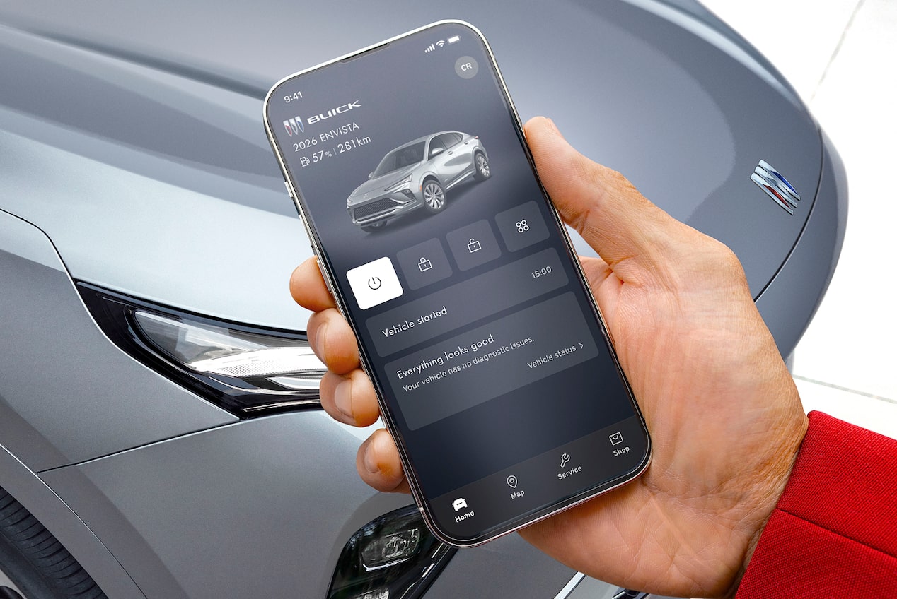 A hand holds a smartphone displaying the Buick mobile app with remote vehicle controls with a grey Envista 2026 in the background of an outdoor setting.