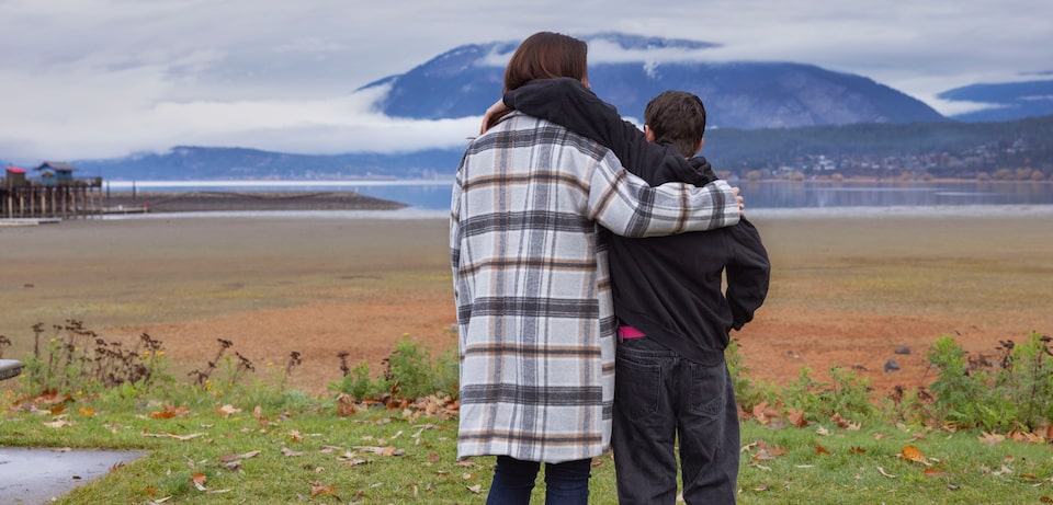 Sarah et son fils, vêtus de manteaux, s'enlacent en contemplant un vaste lac et des montagnes lointaines sous un ciel nuageux.