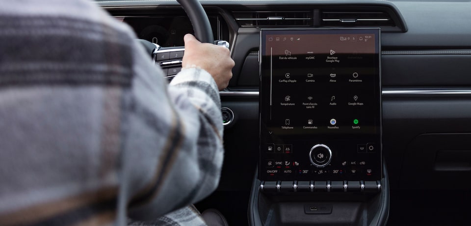 La main d'un conducteur est posée sur le volant, à côté d'un grand écran d'infodivertissement vertical, à l'intérieur d'un GMC Terrain.
